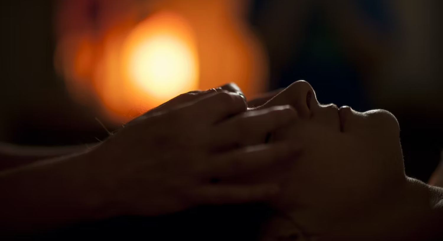 Close-up of a woman's hands massaging the forehead and eyes of another woman, with a calming light in the background.